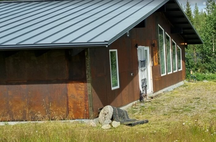 Ett hus med nytt tak och rostiga plåtar på väggarna, med maskering för dörren och stenar, samt spolar som ligger på marken framför.