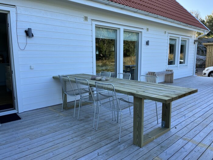 En träbord utomhus framför ett vitt hus med röda takpannor och stora fönster. Bordet är långt och robust med träplankor och åtta metallstolar runt det.