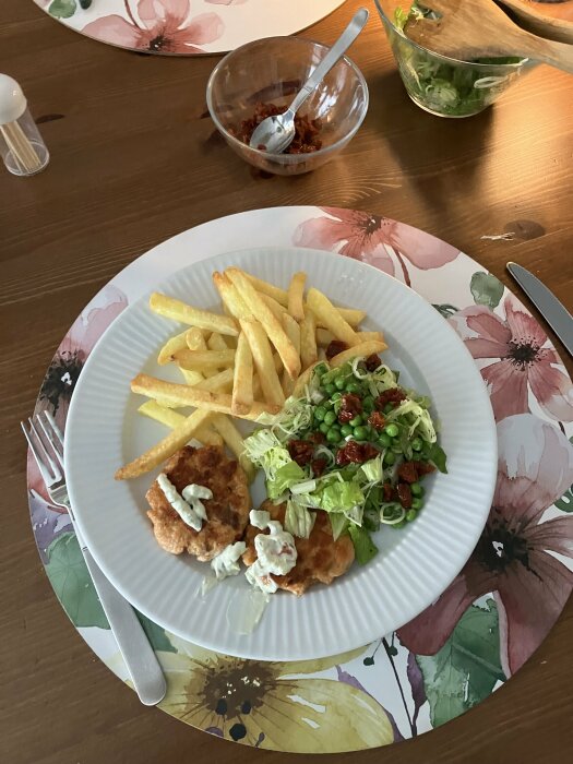 Tallrik med laxbiffar, pommes frites och sallad innehållande bokblad, purjolök, ärtor och soltorkade tomater på ett blommigt underlägg.