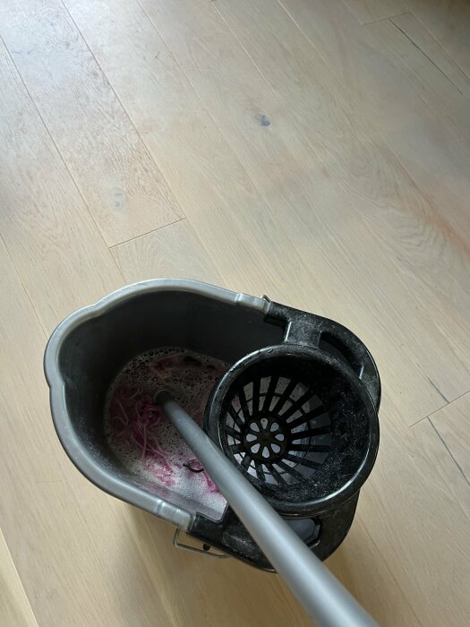 Mopp och hink med rosa vatten står på ett trägolv.