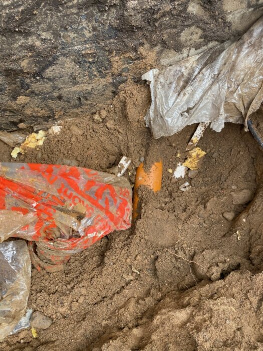 Avgrävt avloppsrör i grävmassor under huset. Plastsäckar och sand synliga runt området. Diskuterar extra kostnader för dränering.