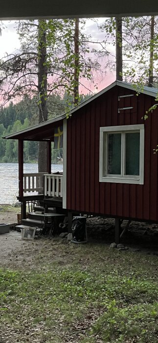 En röd stuga med vita fönster och en veranda, omgiven av träd och sjö i bakgrunden. Marken är täckt av löv och gräs.