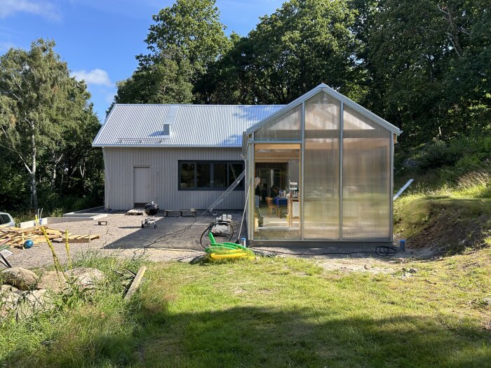 Grått hus med metalltak under konstruktion, med ett uterum med plastväggar till höger. Byggmaterial ligger utspritt på marken framför huset.