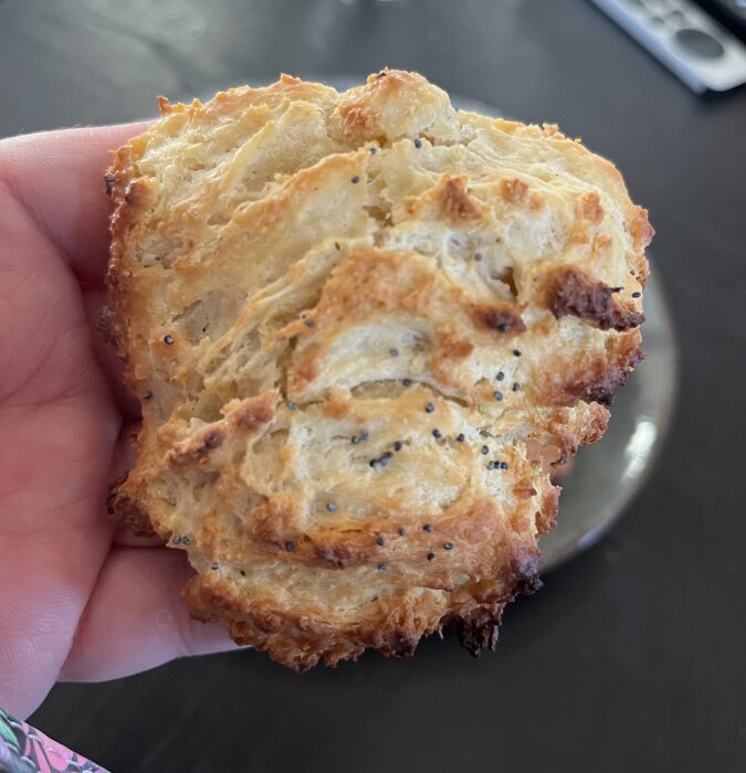 Hand håller ett nybakat scone med en krispig yta, dekorerad med vallmofrön. Sconet har en gyllenbrun färg och visas mot en suddig bakgrund.