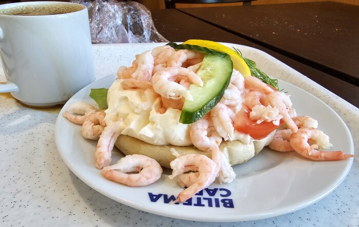 Räksmörgås med majonnäs, skivad gurka, tomat och citron på vitt bröd, serverad på en tallrik märkt "Biltema Café", med en kopp kaffe bredvid.