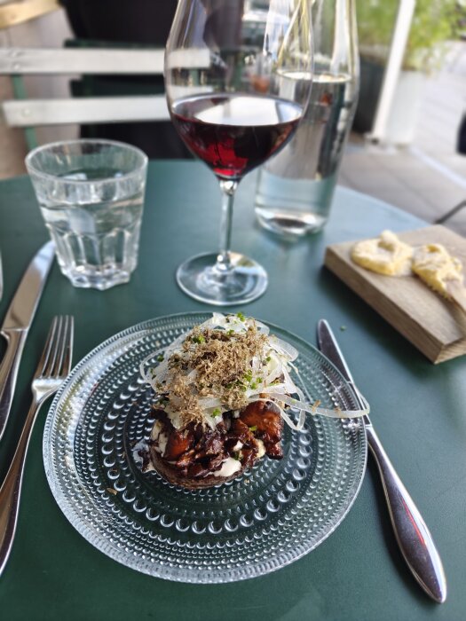 En tallrik med mat, ett glas rödvin, ett glas vatten och ett fat med bröd på ett dukat bord.