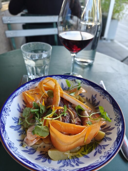 Tallrik med elegant upplagt mat med kött, grönsaker och örter bredvid ett glas rött vin på ett bord. Bakgrunden visar en restaurangmiljö.