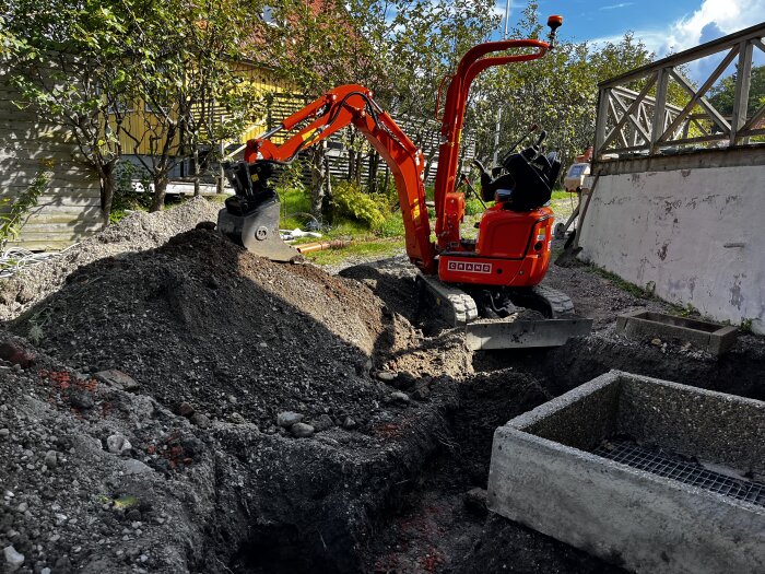 En röd grävmaskin i arbete med att gräva en grop bredvid en vit husgrund i en trädgård. En grävd schakt med grus och en betongbrunn syns i förgrunden.