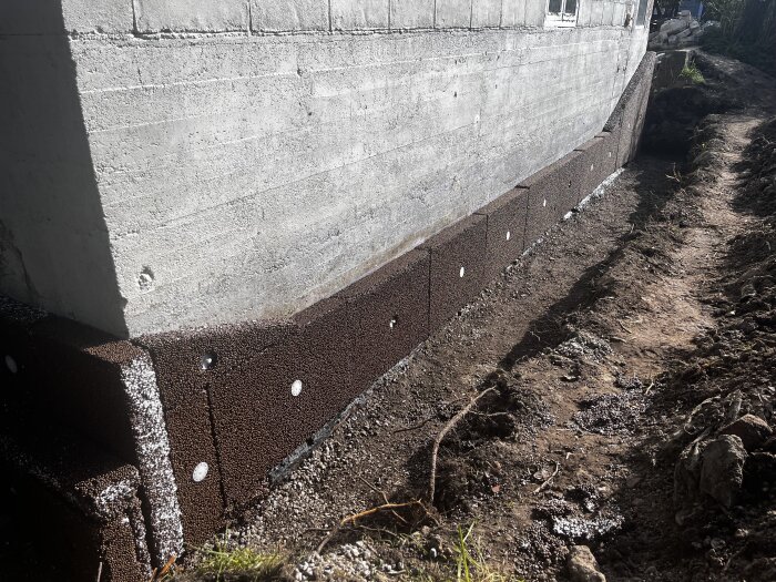 Grundmur med nylagd dränering och isolering, samt grävd schakt runt om. Betongväggen har nyligen fått ett membranapplicerat och dränskivor installerade.