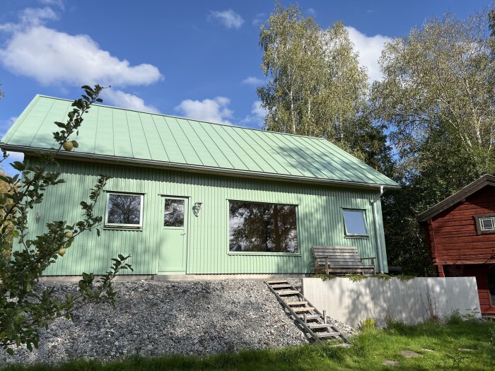 Grönt hus med aluzink-tak och stuprör. En grön dörr och två fönster syns på fasaden. Ett rött hus syns till höger och en träbänk framför huset.