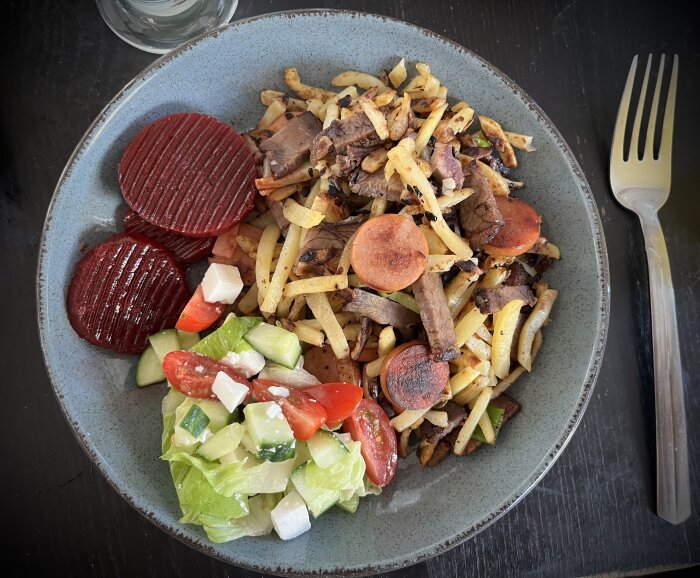 Tallrik med pyttipanna, innehållande potatis, lök, brisket och skivad kabanoss, serverad med sallad och inlagda rödbetor. En gaffel bredvid.