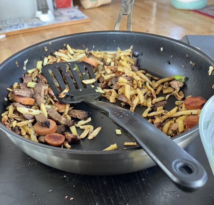 En stekpanna med pyttipanna innehållande potatis, lök, brisket och skivad kabanoss, med stekspade.