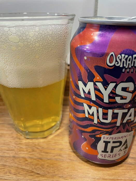 Ölglas fyllt med skummande öl bredvid en burk märkt "Oskar Blues, Myster Mutant, Experimental IPA Series 11" med lila och orange design på ett träbord.