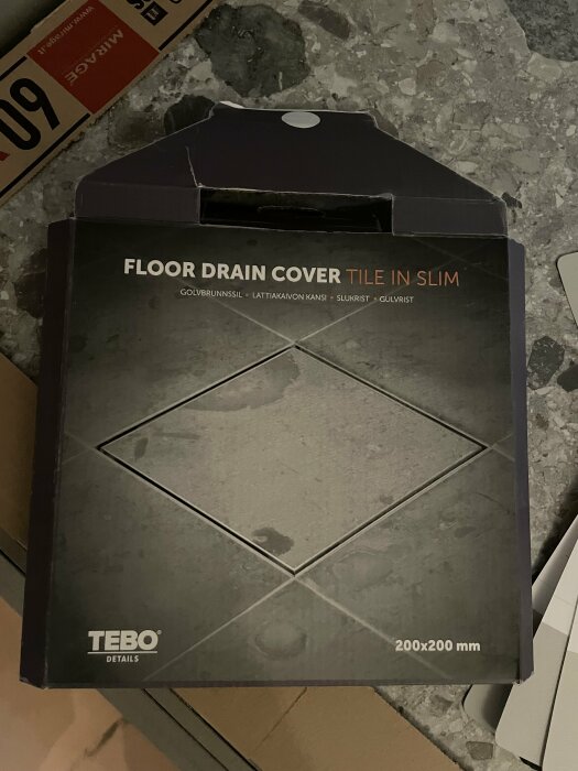 Förpackning för golvbrunnssil av märket TEBO DETAILS med texten "FLOOR DRAIN COVER TILE IN SLIM" på ett betonggolv.