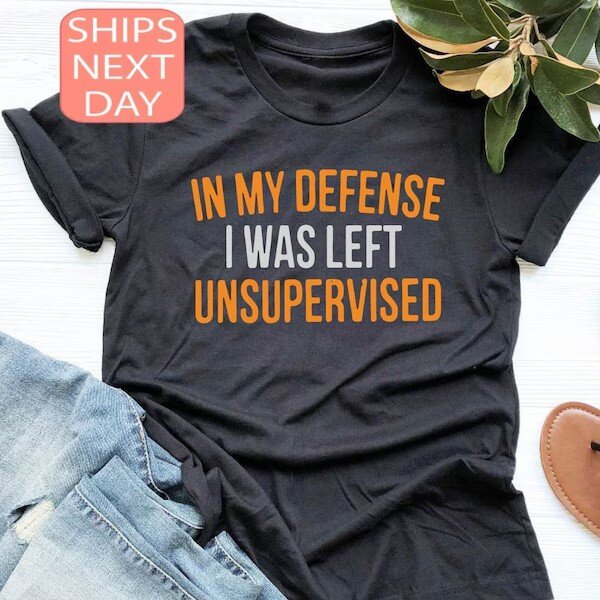 Svart t-shirt med texten "In my defense I was left unsupervised" i orange och grått, bredvid ett par blå jeans.