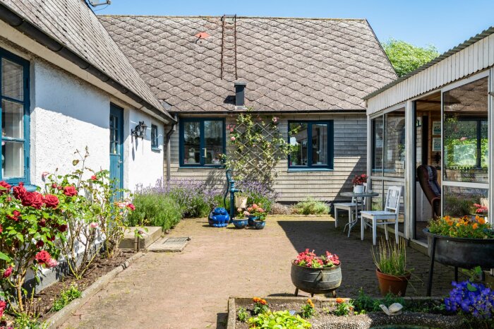 Villa med trädgård och uteplats, vitmålade väggar blandat med träpanel, blå fönster och dörrar, omgiven av blommor och buskar, mot en klarblå himmel.