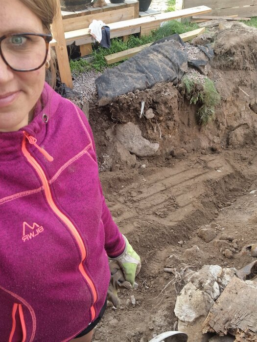 Kvinna i rosa jacka står bredvid en utgrävd schakt där altanen har rivits för att byta badrumsstammar. Diverse bråte och trä syns runt utgrävningen.