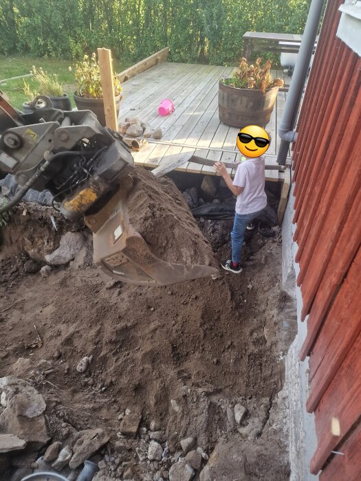 Grävmaskin och person vid pågående arbete med att byta badrumsstammar vid grunden av ett rött hus, där en altan delvis demonterats.