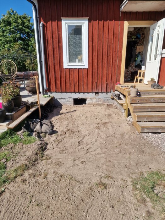 Röd träfasad med vitt fönster och öppen dörr till ett hus. Utrymme framför där altanen rivits och ett runt hål sågat i grunden, marken täckt med jord.