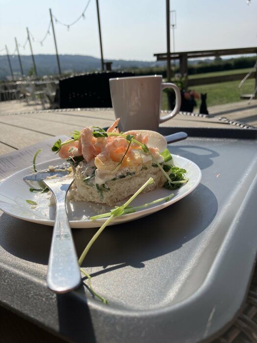 Räksmörgås på en tallrik med bestick, en kaffekopp, utomhus på ett kafé, med utsikt över en äng och avlägsna kullar.