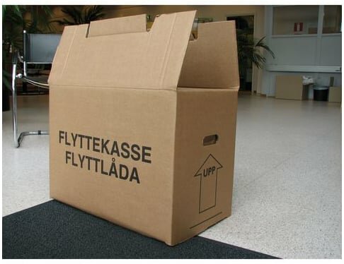 Flyttkartong i wellpapp med texten "FLYTTEKASSE FLYTTLÅDA" står öppen i ett inomhusutrymme med ljus golvyta.