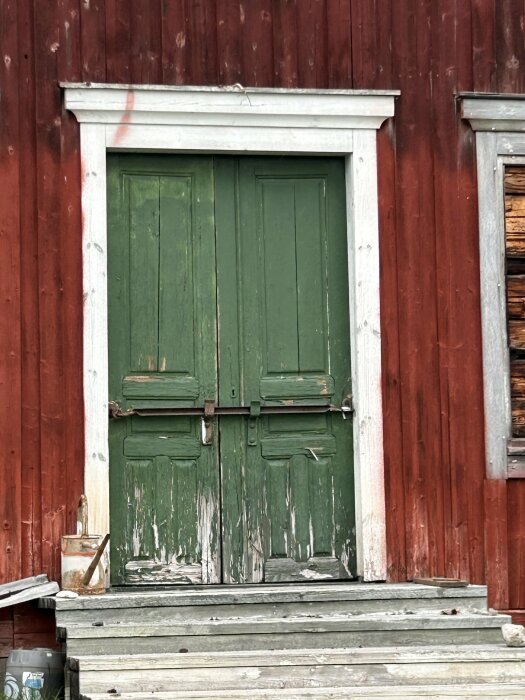 Slitna gröna dörrar på ett rött trähus, med flagnande målarfärg och en pensel och färgburk på trappan utanför.