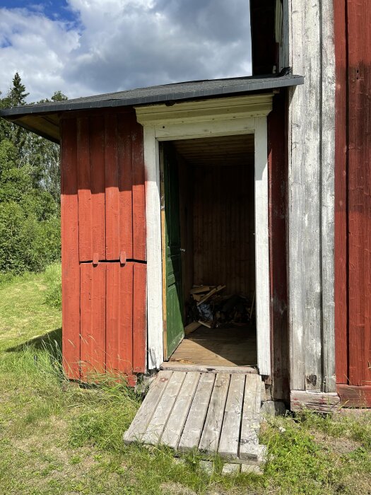 Röd stuga med en öppen grön dörr och ramp, omgiven av grönska. Solen skiner och himlen är delvis molnig.