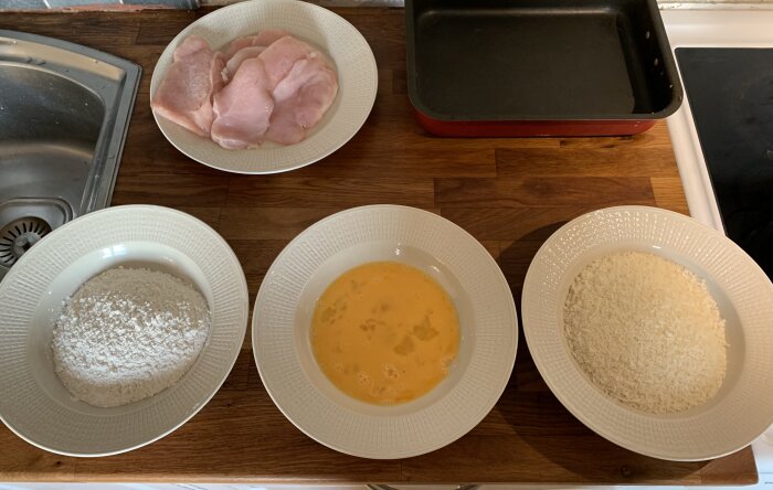 Ingredienser för att panera kyckling ligger i skålar på en diskbänk: mjöl, ägg och ströbröd, med rå kycklingbröstskivor och en plåt i närheten.