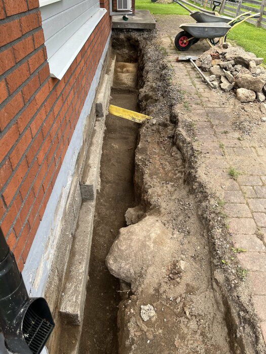 Grävd grop längs husgrund av tegel med synliga lecablock och sand, en skottkärra med sten och verktyg syns i bakgrunden på stenen.