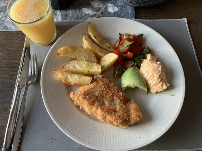 Tallrik med panerad fisk, ugnsbakad potatis, avokado, sågade tomater, krämig dressing, och ett glas apelsinjuice. Bestick på en bordsmatta bredvid tallriken.