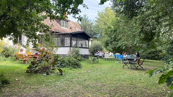 Uteserveringen på Olof Viktors kafé med gäster som sitter vid bord i trädgården, omgiven av gröna träd och buskar, med en byggnad i bakgrunden.