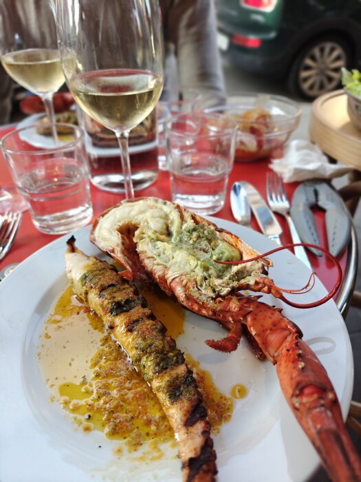 Grillad hummer och annan skaldjursrätt på en tallrik, omgiven av vinglas och glas med vatten på ett dukat bord utomhus.