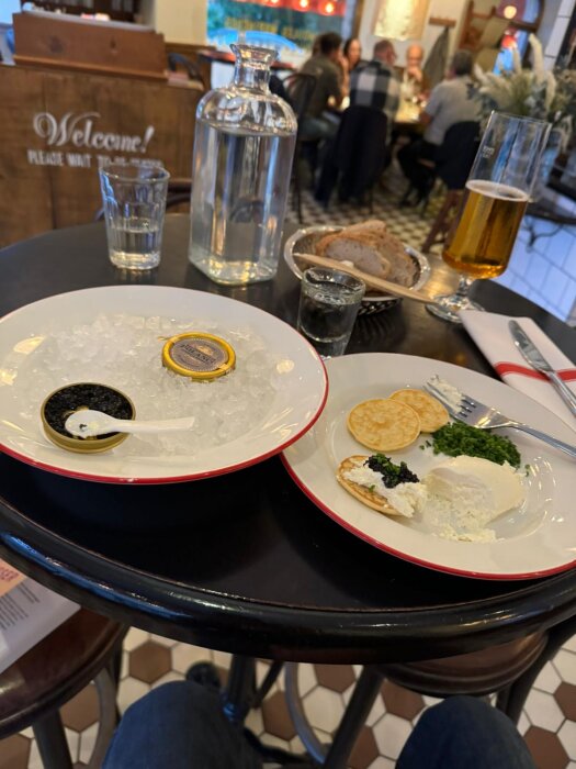 Caviar och tillbehör serverade på en restaurangbord med drycker och bröd i bakgrunden. En plastsked ligger i caviaren.