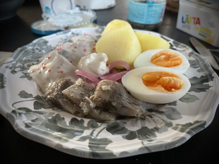 Silltallrik med brantevikssill och skärgårdssill, kokt potatis, kokt ägg, gräddfil, picklad rödlök och en ostmacka på en blommig tallrik.