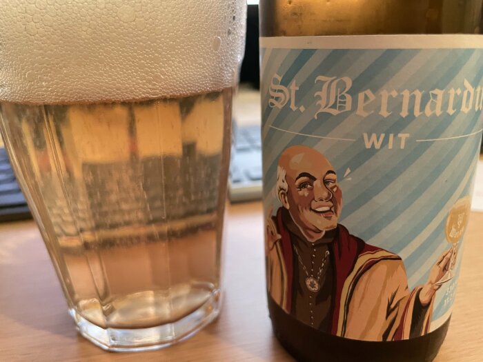 Ett glas med skummande öl bredvid en flaska med etiketten "St. Bernardus Wit" och en illustration av en leende munk.