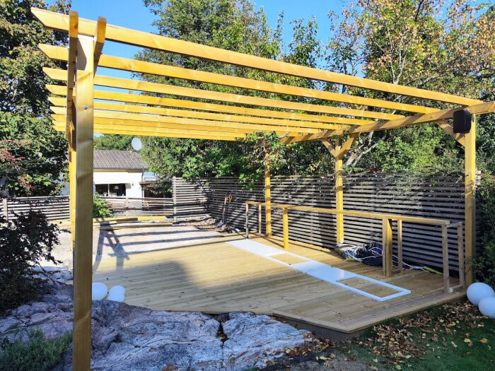 Trädäck med pergola färdigbyggd, klart för steg två i byggandet av utekök; markerad plats för köksdelar, omgiven av grönska.