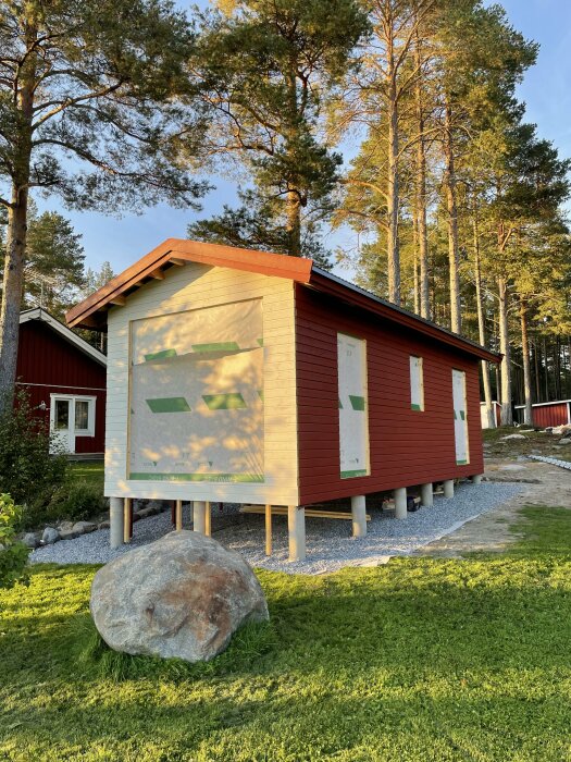 Ett nästan färdigmålat hus på pålar vid trädgränsen, med en ofärdig fasad och ett stort stenblock i förgrunden.