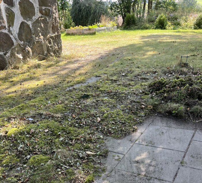 Stenlagd yta vid sidan av ett stenhus delvis täckt av mossa och växtlighet, omgiven av en trädgård med buskar och högt gräs i bakgrunden.