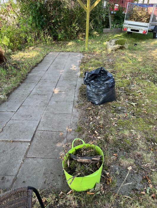 En nyligen framröjd stenläggning bredvid ett hus, med en svart sopsäck och en grön hink fylld med växtrester i fokus.