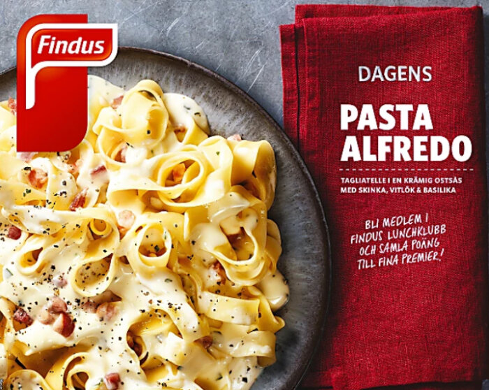 Findus förpackning av dagens pasta Alfredo, tagliatelle i krämig ostsås med skinka, vitlök och basilika, mot en grå bakgrund och röd servett med marknadsföringstext.