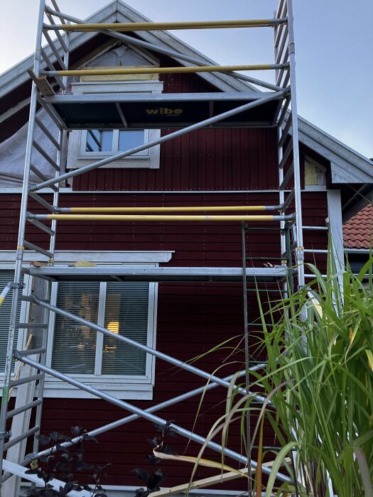 Byggnadsställning framför en röd fasad med vita fönster och foder, första våningen visar ett fönster. Arbetsplattformen når till fönstret på andra våningen.