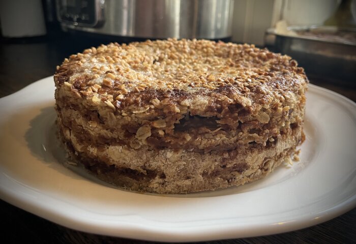 Havrekaka med lager av rivna äpplen och torra ingredienser, toppad med hackad mandel, presenterad på en vit tallrik.