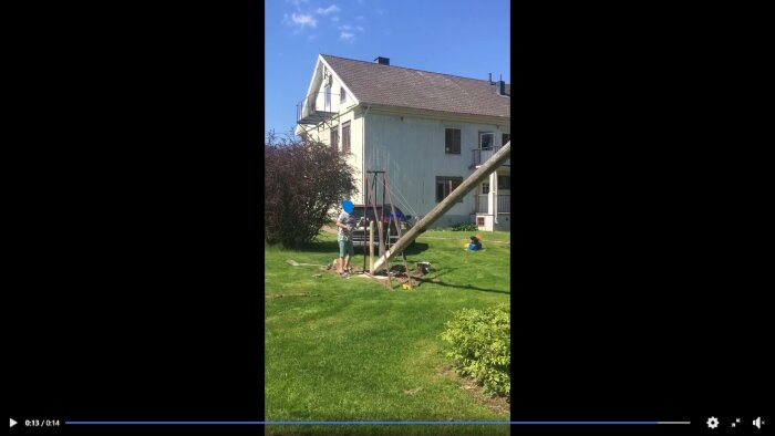 Man använder en fällanordning för att sänka en flaggstång bredvid ett hus i en trädgård. Ett barn sitter på gräsmattan.