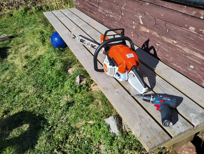 En orange Stihl motorsåg och en Bosch skruvdragare ligger på en bänk gjord av trallbrädor utomhus nära en trävägg.