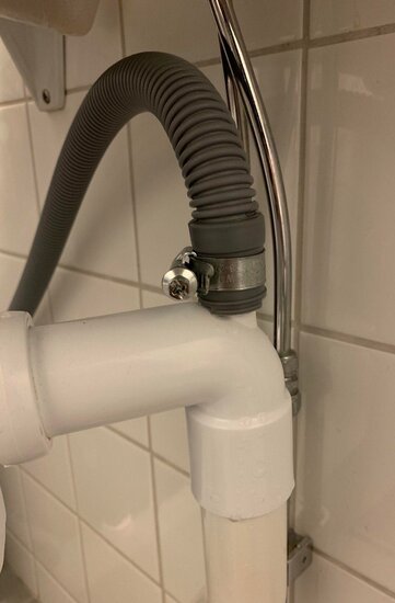 Installation av grå dräneringsslang fäst med slangklämma på ett vitt PVC-avlopp mot vit kakelvägg.