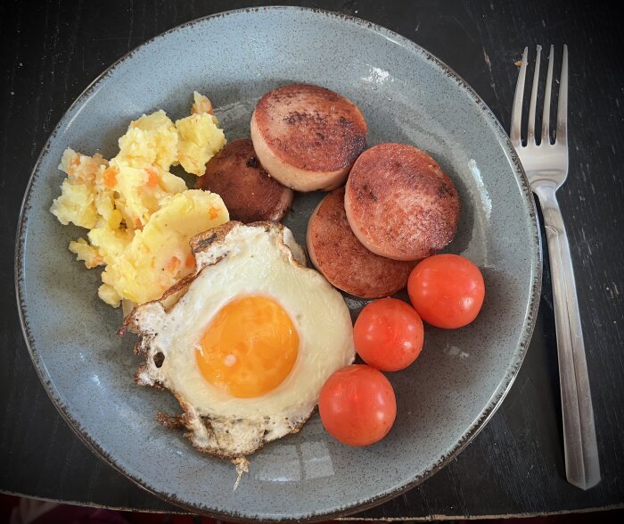 En tallrik med stekt falukorv, ett stekt ägg sunny side up, potatismos och tre små tomater. En gaffel ligger vid sidan av tallriken.