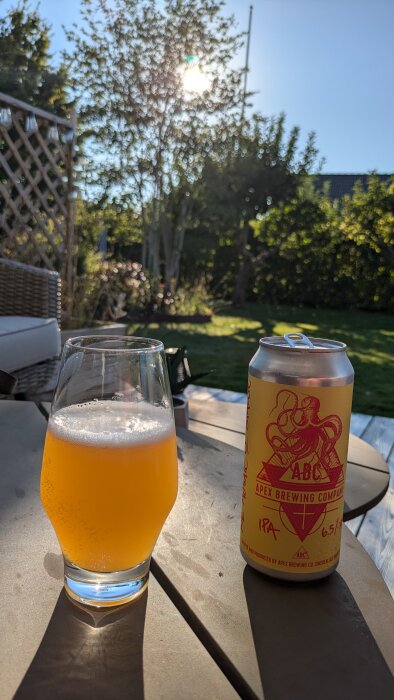 En burk Apex Brewing Company IPA och ett halvfullt glas öl på en uteplats i sommarträdgård med solstrålar i bakgrunden.