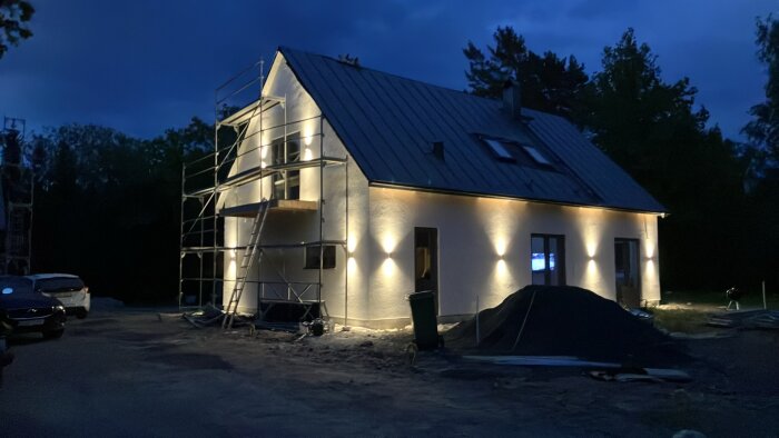 Ett hus under renovering i kvällsbelysning, med byggnadsställningar runt fasaden, upplysta ytterväggar och bilar parkerade framför.