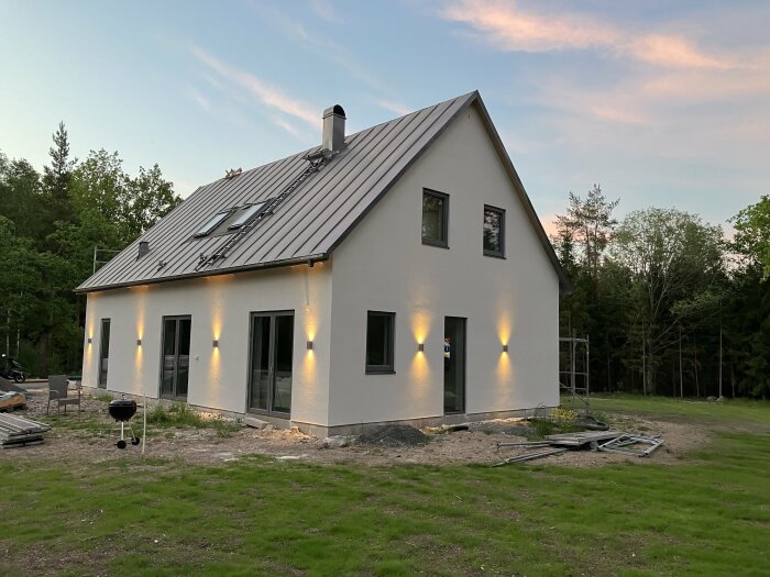 Ett nybyggt hus med vit fasad och mörkt tak, utvändiga lampor på väggen och trädgård som fortfarande anläggs. Träd och skog i bakgrunden.