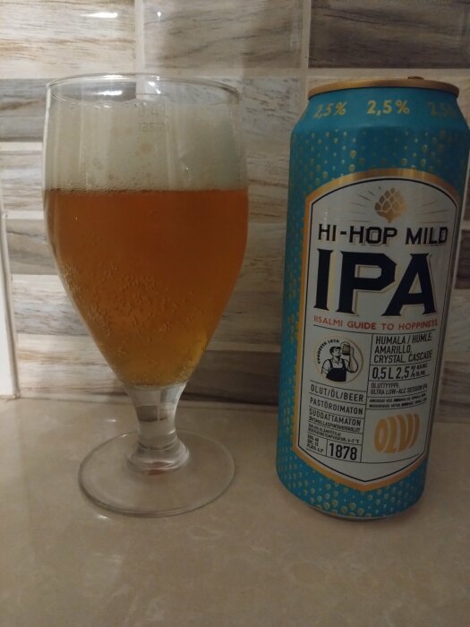 Ett glas med öl bredvid en burk Olvi Hi-Hop Mild IPA 2,5% på ett köksbänk, med klinkers i bakgrunden.